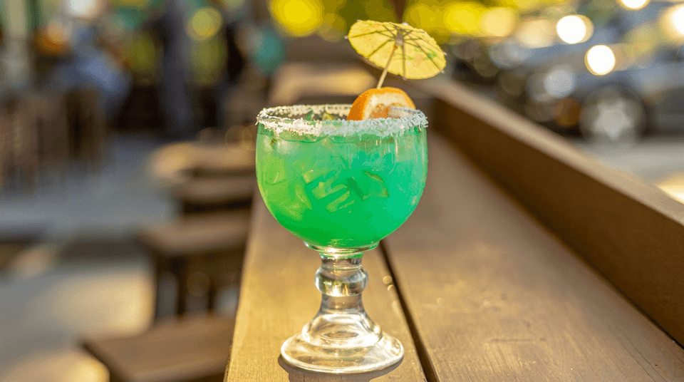 Bright green cocktail in a salted-rim goblet, garnished with orange and a paper umbrella on a warmly lit restaurant bar.