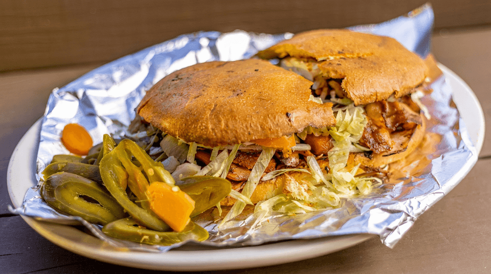Two loaded burgers with lettuce, tomatoes, onions, and bacon sit on foil alongside sliced pickled jalapeños and carrots.